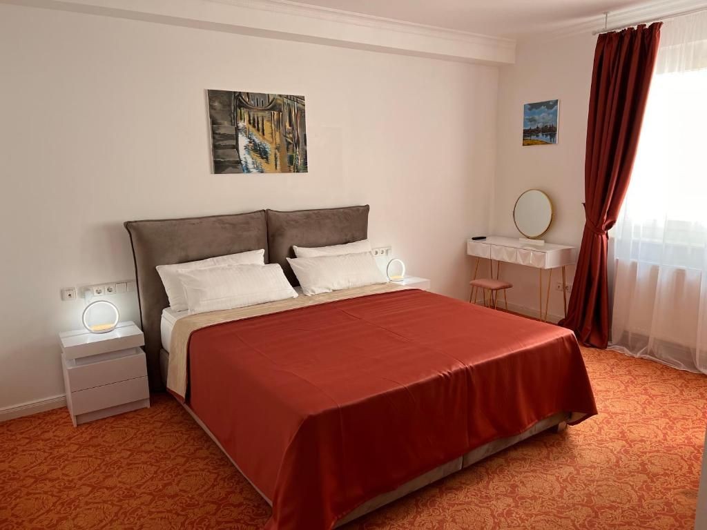 Helles Hotelzimmer mit großem Doppelbett, roter Bettdecke, zwei Nachttischen mit runden Lampen, Schminktisch mit Spiegel und Hocker sowie bodenlangen Vorhängen vor einem großen Fenster.