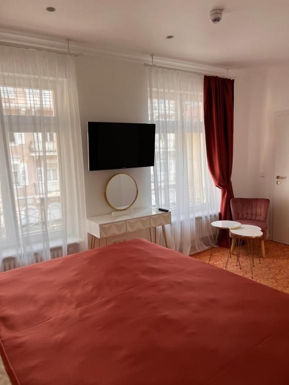 Helles Hotelzimmer mit bodentiefen Fenstern, Flachbild-TV, Schminktisch mit Spiegel, gemütlichem Sessel und Beistelltisch – kombiniert mit roten Vorhängen und passender Bettdecke.