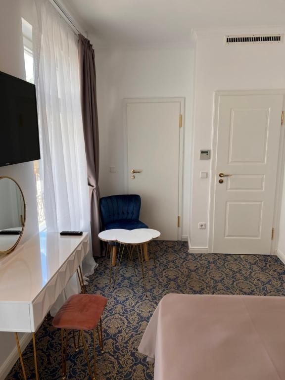 Teilansicht des Zimmers „Kurhaus“ im Art Hotel House One in Baden-Baden mit floralen Teppichboden, Schminktisch mit Spiegel, rosa Hocker, TV, blauer Sessel, weißen Beistelltischen und zwei Zimmertüren.